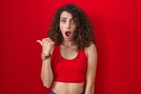 Photo for Hispanic woman with curly hair standing over red background surprised pointing with hand finger to the side, open mouth amazed expression. - Royalty Free Image