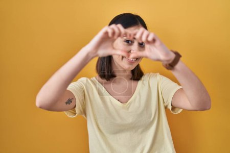 Photo for Hispanic girl wearing casual t shirt over yellow background doing heart shape with hand and fingers smiling looking through sign - Royalty Free Image