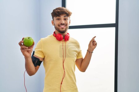 Photo for Arab man with beard wearing sportswear eating green apple smiling happy pointing with hand and finger to the side - Royalty Free Image