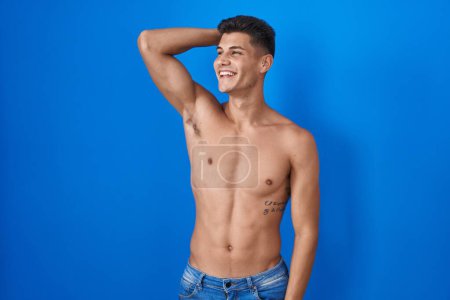 Téléchargez les photos : Jeune homme hispanique debout torse nu sur fond bleu souriant cheveux touchants confiants avec geste de la main vers le haut, posant attrayant et à la mode - en image libre de droit