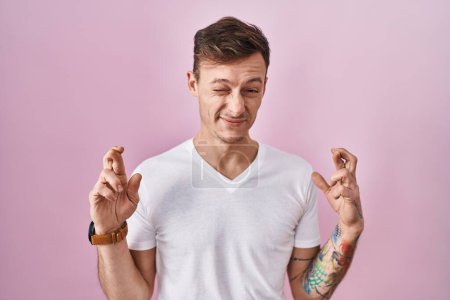 Téléchargez les photos : Homme caucasien debout sur fond rose geste doigt croisé souriant avec espoir et les yeux fermés. chance et concept superstitieux. - en image libre de droit