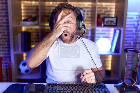 Foto de Hombre de mediana edad con barba jugando videojuegos con auriculares asomándose en shock cubriendo la cara y los ojos con la mano, mirando a través de los dedos con expresión avergonzada. - Imagen libre de derechos