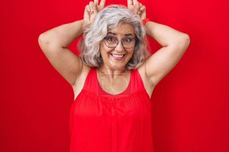 Foto de Mujer de mediana edad con el pelo gris de pie sobre fondo rojo posando divertido y loco con los dedos en la cabeza como orejas de conejo, sonriente alegre - Imagen libre de derechos