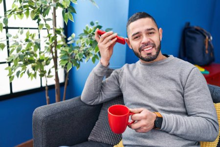 Téléchargez les photos : Jeune homme latino écoute un message vocal par smartphone boire du café à la maison - en image libre de droit