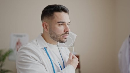 Photo for Young hispanic man doctor standing with serious expression taking out medical mask at clinic - Royalty Free Image