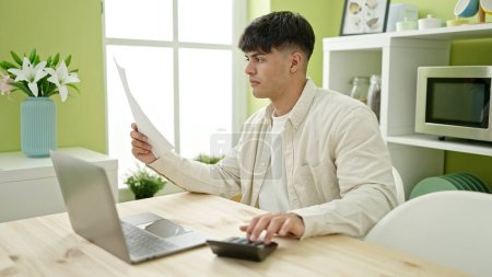 Téléchargez les photos : Jeune homme hispanique utilisant ordinateur portable et calculatrice assis sur la table à manger - en image libre de droit