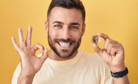 Foto de Hombre hispano guapo sosteniendo moneda criptomoneda tron haciendo signo ok con los dedos, sonriendo gesto amistoso excelente símbolo - Imagen libre de derechos