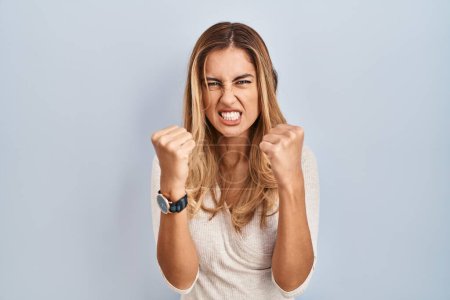 Photo for Young blonde woman standing over isolated background angry and mad raising fists frustrated and furious while shouting with anger. rage and aggressive concept. - Royalty Free Image