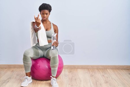 Photo for African american woman wearing sportswear sitting on pilates ball showing middle finger, impolite and rude fuck off expression - Royalty Free Image