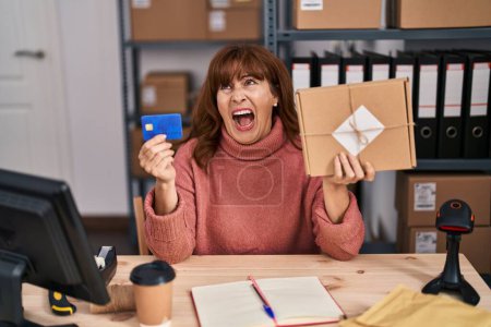 Foto de Mujer hispana de mediana edad que trabaja en el comercio electrónico de pequeñas empresas con tarjeta de crédito enojada y loca gritando frustrada y furiosa, gritando con ira mirando hacia arriba. - Imagen libre de derechos