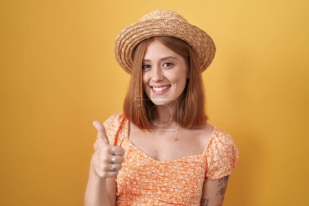 Téléchargez les photos : Jeune femme rousse debout sur fond jaune portant un chapeau d'été faisant des pouces heureux geste avec la main. approuvant l'expression regardant la caméra montrant le succès. - en image libre de droit