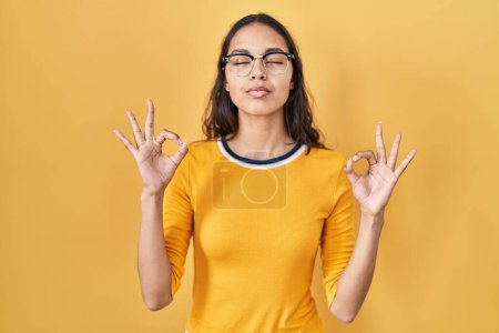 Téléchargez les photos : Jeune femme brésilienne portant des lunettes sur fond jaune se détendre et sourire les yeux fermés en faisant geste de méditation avec les doigts. concept de yoga. - en image libre de droit