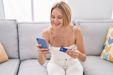 Téléchargez les photos : Jeune femme blonde utilisant smartphone et carte de crédit assis sur le canapé à la maison - en image libre de droit
