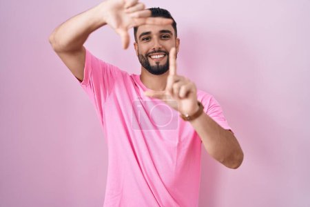 Téléchargez les photos : Jeune homme hispanique debout sur fond rose souriant faisant cadre avec les mains et les doigts avec le visage heureux. concept de créativité et photographie. - en image libre de droit