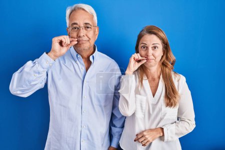 Téléchargez les photos : Couple hispanique du moyen âge debout sur fond bleu bouche et lèvres fermées comme zip avec les doigts. secret et silencieux, tabou parlant - en image libre de droit