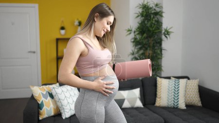 Photo for Young pregnant woman holding yoga mat touching belly at home - Royalty Free Image