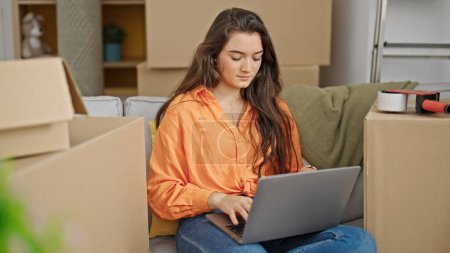 Photo for Young beautiful hispanic woman using laptop sitting on sofa at new home - Royalty Free Image