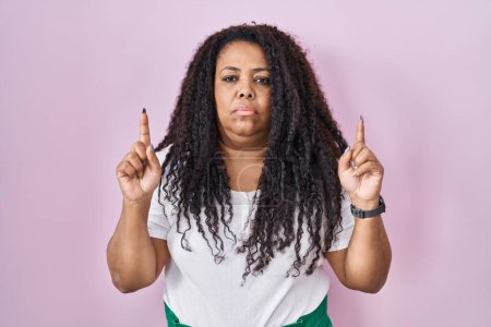 Photo for Plus size hispanic woman standing over pink background pointing up looking sad and upset, indicating direction with fingers, unhappy and depressed. - Royalty Free Image