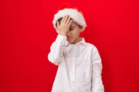 Téléchargez les photos : Petit garçon caucasien portant chapeau de Noël sur fond rouge surpris avec la main sur la tête pour erreur, rappelez-vous erreur. oublié, mauvais concept de mémoire. - en image libre de droit