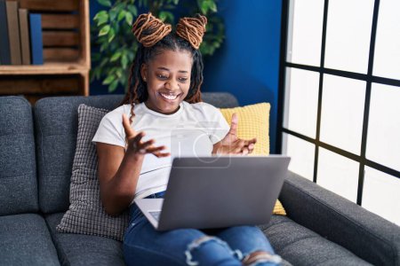 Foto de Mujer africana con el pelo trenzado usando portátil en casa mirando a la cámara sonriendo con los brazos abiertos para abrazar. expresión alegre abrazando la felicidad. - Imagen libre de derechos