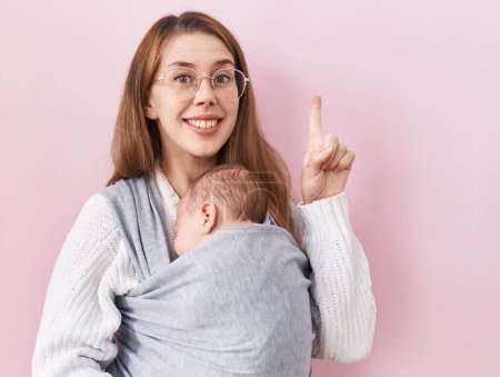 Foto de Joven mujer caucásica sosteniendo y llevando al bebé en un cabestrillo sorprendido con una idea o pregunta señalando el dedo con la cara feliz, número uno - Imagen libre de derechos