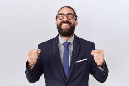 Téléchargez les photos : Homme hispanique avec barbe en costume et cravate excité pour le succès avec les bras levés et les yeux fermés célébrant la victoire en souriant. concept gagnant. - en image libre de droit