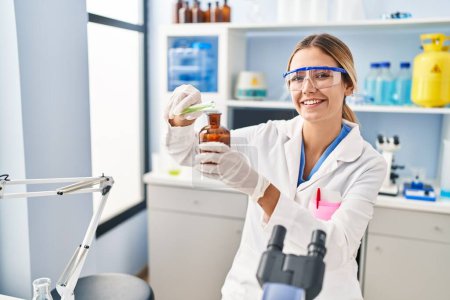 Photo for Young hispanic woman scientist pouring liquid on bottle at laboratory - Royalty Free Image