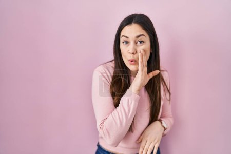 Téléchargez les photos : Jeune femme brune debout sur fond rose main sur bouche racontant rumeur secrète, chuchotant conversation malicieuse - en image libre de droit