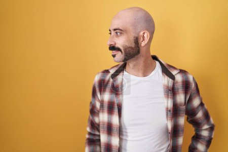 Foto de Hombre hispano con barba de pie sobre fondo amarillo mirando hacia otro lado con sonrisa en la cara, expresión natural. riendo confiado. - Imagen libre de derechos