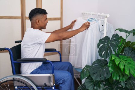 Foto de Hombre latino joven eligiendo camisa sentada en silla de ruedas en el dormitorio - Imagen libre de derechos
