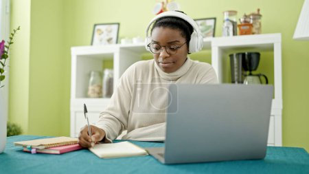 Téléchargez les photos : Femme afro-américaine utilisant un ordinateur portable et écouteurs écrivant des notes étudier à la salle à manger - en image libre de droit
