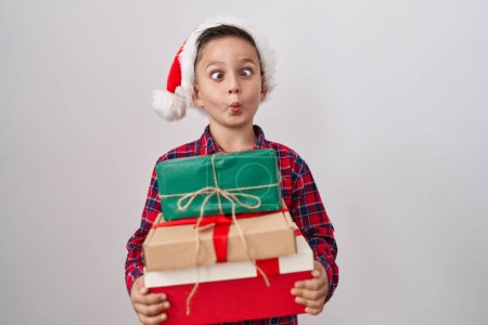 Foto de Pequeño niño hispano con sombrero de navidad sosteniendo regalos haciendo cara de pez con boca y ojos entrecerrados, loco y cómico. - Imagen libre de derechos
