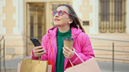 Foto de Mujer hispana madura con el pelo gris sonriendo yendo de compras usando smartphone en la calle - Imagen libre de derechos