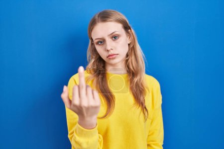 Foto de Young caucasian woman standing over blue background showing middle finger, impolite and rude fuck off expression - Imagen libre de derechos