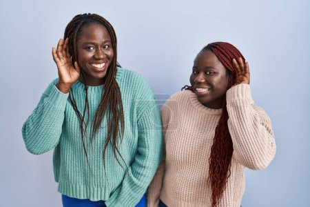 Foto de Dos mujeres africanas de pie sobre fondo azul sonriendo con la mano sobre el oído escuchando un rumor o chismes. concepto de sordera. - Imagen libre de derechos
