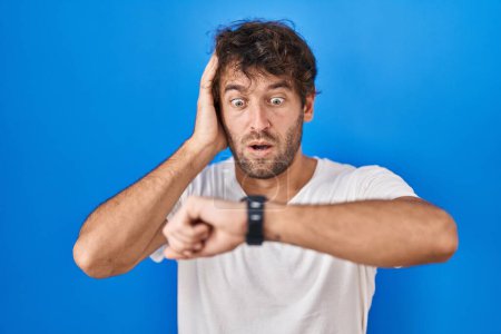 Téléchargez les photos : Jeune homme hispanique debout sur fond bleu regardant le temps de la montre inquiet, peur de se faire tard - en image libre de droit