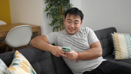 Foto de Hombre chino joven usando teléfono inteligente sentado en el sofá en casa - Imagen libre de derechos