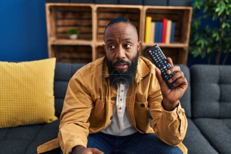 Foto de Hombre afroamericano sosteniendo control remoto de televisión asustado y sorprendido con la boca abierta para sorpresa, cara de incredulidad - Imagen libre de derechos