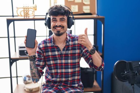 Photo for Young hispanic man with beard showing smartphone screen at music studio smiling happy and positive, thumb up doing excellent and approval sign - Royalty Free Image