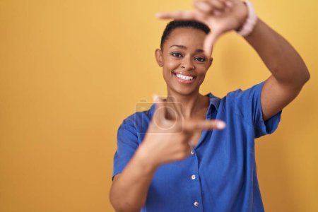 Téléchargez les photos : Femme afro-américaine debout sur fond jaune souriant faisant cadre avec les mains et les doigts avec le visage heureux. concept de créativité et photographie. - en image libre de droit