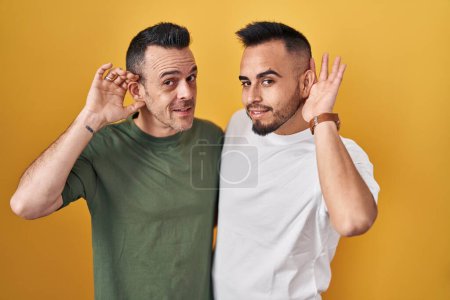 Téléchargez les photos : Couple homosexuel debout sur fond jaune souriant avec la main sur l'oreille à l'écoute d'une audience à la rumeur ou des commérages. concept de surdité. - en image libre de droit