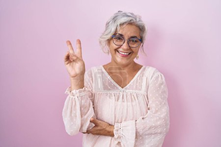 Téléchargez les photos : Femme d'âge moyen avec les cheveux gris debout sur fond rose souriant avec le visage heureux clin d'oeil à la caméra faisant signe de victoire. numéro deux. - en image libre de droit