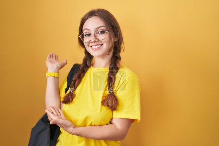 Foto de Joven mujer caucásica con mochila de estudiante sobre fondo amarillo invitando a entrar sonriendo natural con la mano abierta - Imagen libre de derechos