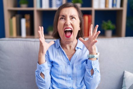 Photo for Middle age hispanic woman sitting on the sofa at home crazy and mad shouting and yelling with aggressive expression and arms raised. frustration concept. - Royalty Free Image