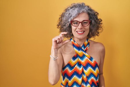Téléchargez les photos : Femme d'âge moyen avec les cheveux gris debout sur fond jaune geste souriant et confiant avec la main faisant signe de petite taille avec les doigts regardant et la caméra. concept de mesure. - en image libre de droit