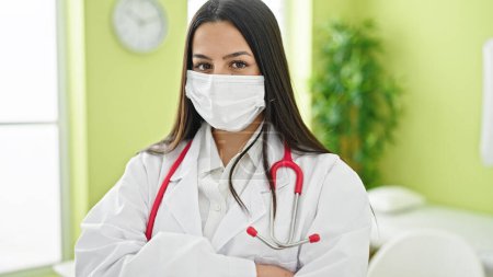 Foto de Joven mujer hispana hermosa médico de pie con los brazos cruzados gesto usando máscara médica en la clínica - Imagen libre de derechos