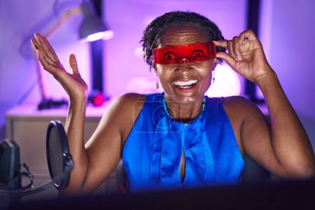 Téléchargez les photos : Femme africaine avec dreadlocks jouer à des jeux vidéo portant des lunettes de réalité virtuelle célébrant la réalisation avec sourire heureux et expression gagnante avec la main levée - en image libre de droit