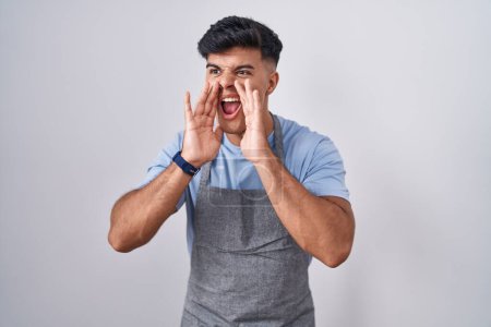 Téléchargez les photos : Jeune homme hispanique portant tablier sur fond blanc criant en colère à haute voix avec les mains sur la bouche - en image libre de droit