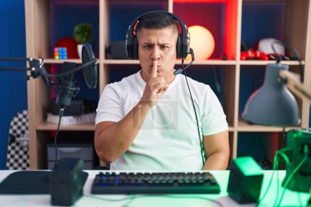 Photo for Young hispanic man playing video games asking to be quiet with finger on lips. silence and secret concept. - Royalty Free Image