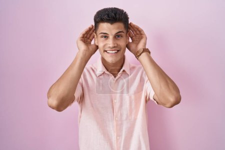 Téléchargez les photos : Jeune homme hispanique debout sur fond rose essayant d'entendre les deux mains sur le geste de l'oreille, curieux de commérages. problème auditif, sourd - en image libre de droit